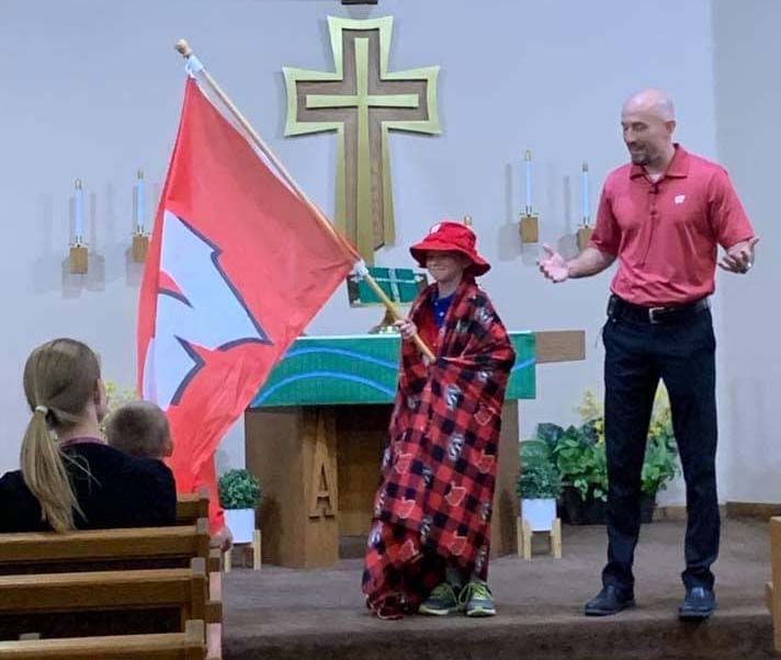Pastor Collin Vanderhoof giving chapel at Pilgrim, Menomonee Falls, Wis. Child shown dressed up and holding University of Wisconsin--Madison flag.