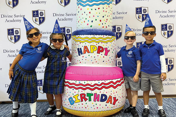 toddlers next to a blow up birthday cake and wearing hats