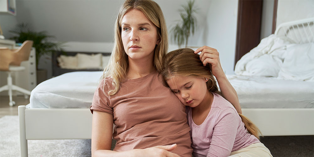 Mom sitting with daughter on the floor of a bedroom playing with daughters hair but looking defeated