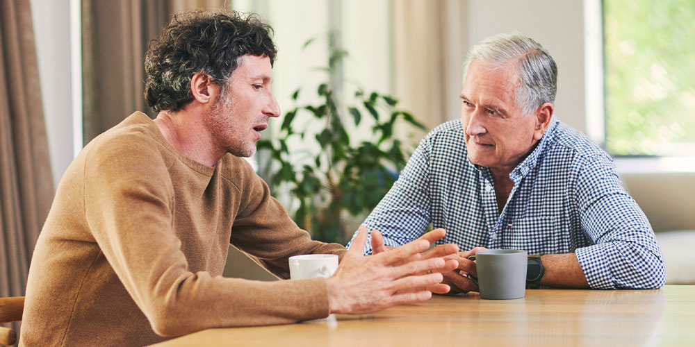 man in tan sweater talking to older man in a blue and white checkered button up shirt
