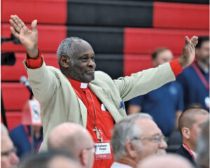 Kanya, pastor in red shirt with arms outstreched.