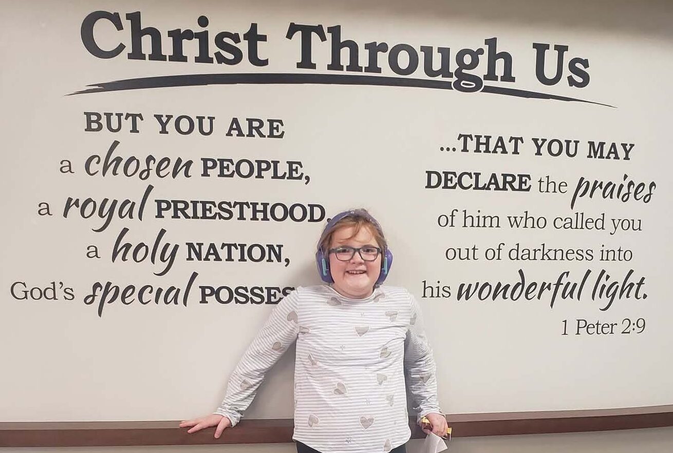 Aria Welke standing in front of a "Christ through us" sign with the words of 1 Peter 2:9 underneath