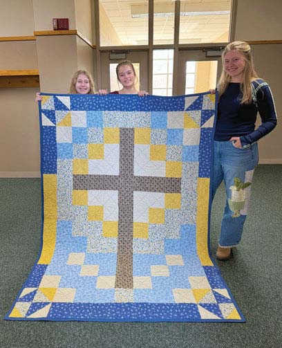 three teen girls holding up quilt with cross on it