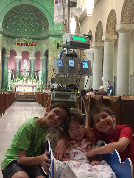 Children Eli, Eliana, and Isaiah Shah in a hospital chapel. Eliana is in a portable hospital bed hooked up to monitors.