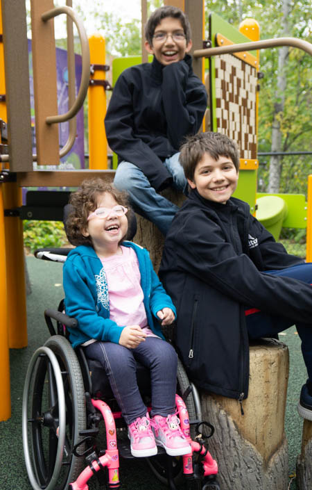 Children Eli, Eliana, and Isaiah Shah laughing in a park. Eliana is in a wheelchair. The two boys are sitting on playground equipment.