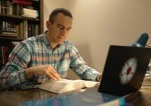 Man at desk looking through Bible studying. Bookcase. computer